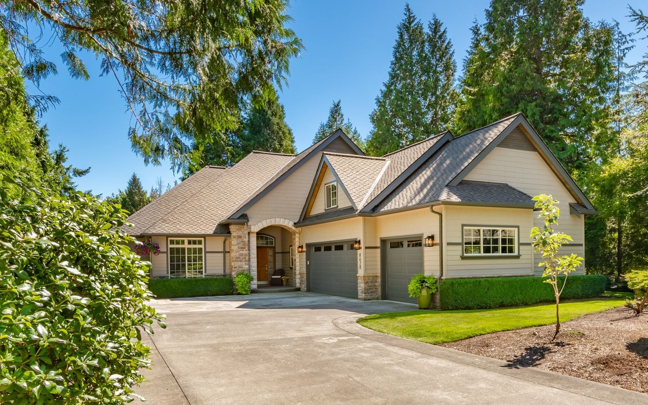 Beige multi-gabled house with stone accents, two-car garage, and a wide driveway amid mature trees.