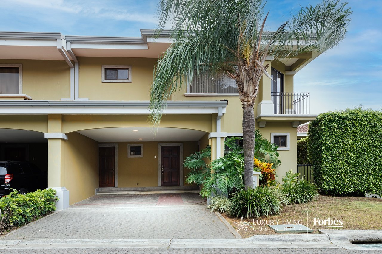 Two-story yellow home with a covered carport, upper balcony, and palm-lined garden.