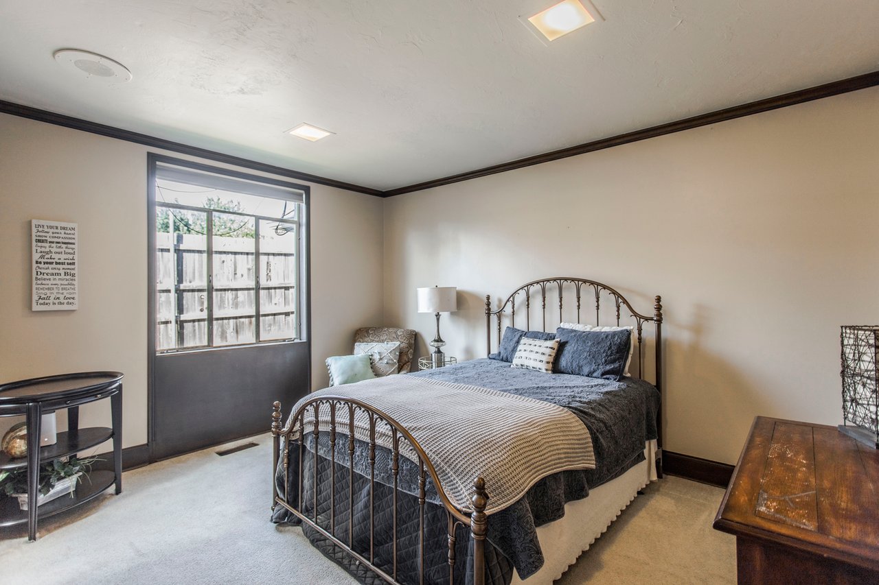 Furnished bedroom with a metal bed, blue-gray bedding, and a large window facing a fenced yard.