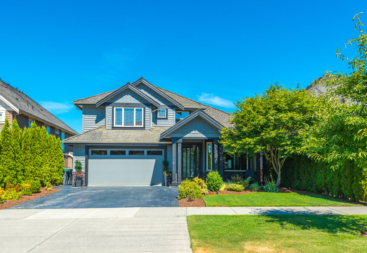 Two-story blue-gray house with a wide garage, landscaped lawn, and mature trees.