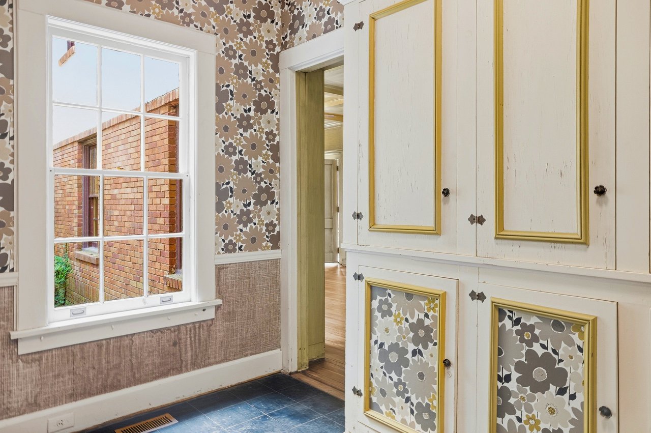 Room with floral wallpaper, white built-in cabinets with gold trim, and a large window.