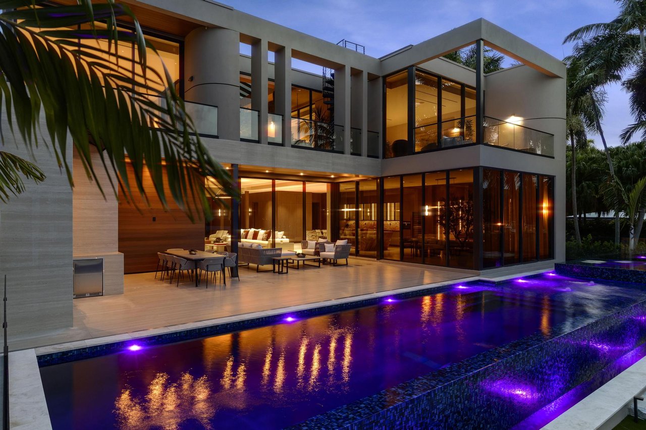 Modern glass-and-concrete home with a purple-lit pool and outdoor dining terrace at dusk.