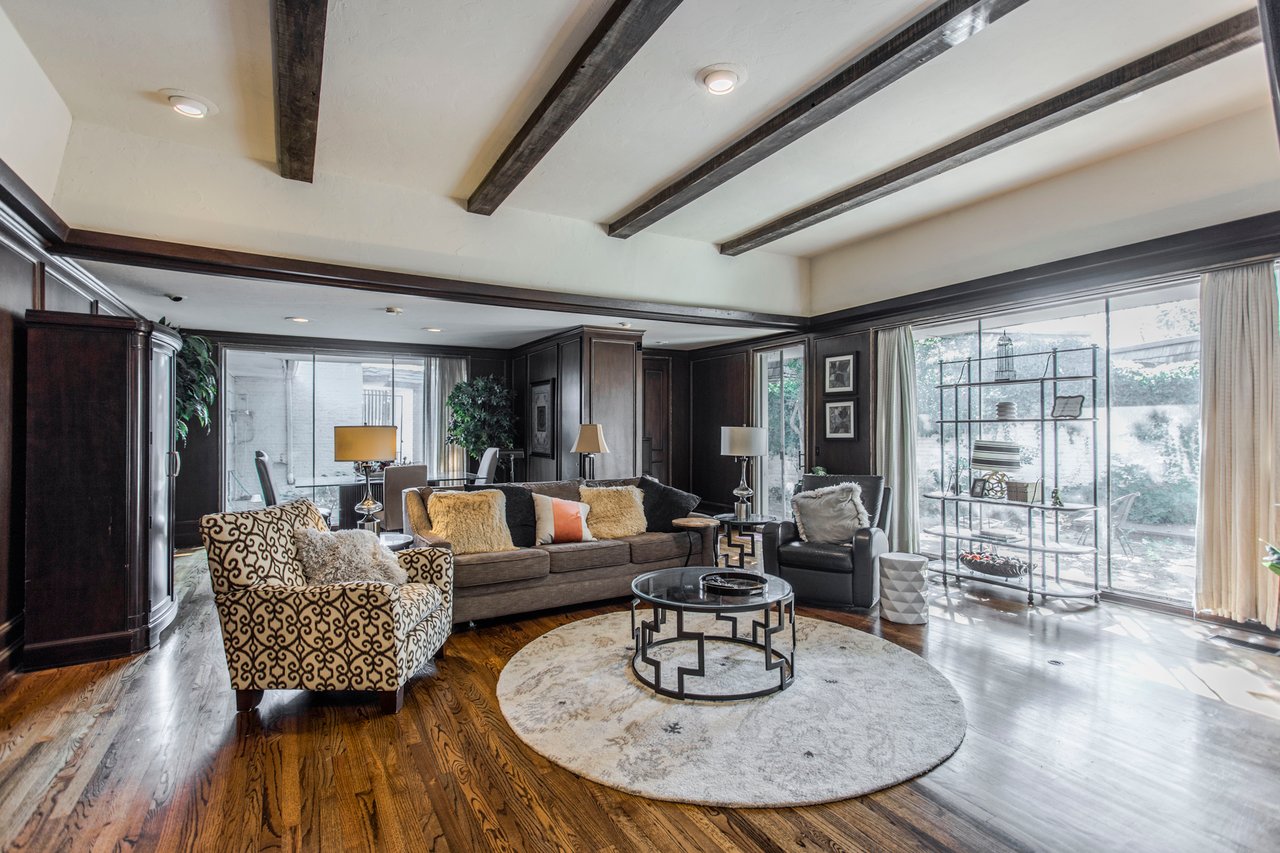 Living room with dark wood paneling, exposed beams, hardwood floors, and large garden-facing windows.
