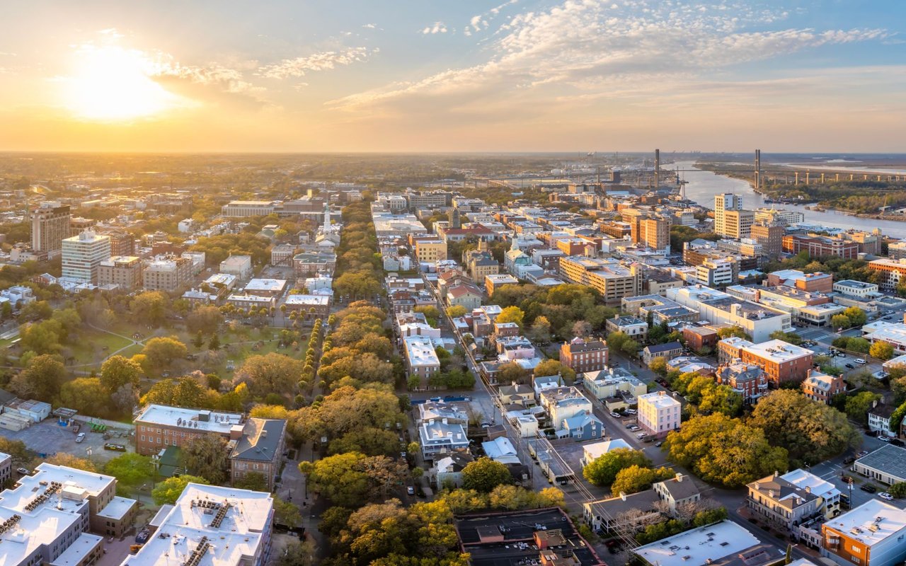 Aerial cityscape beside a broad river, with tree-lined streets glowing in the setting sun.