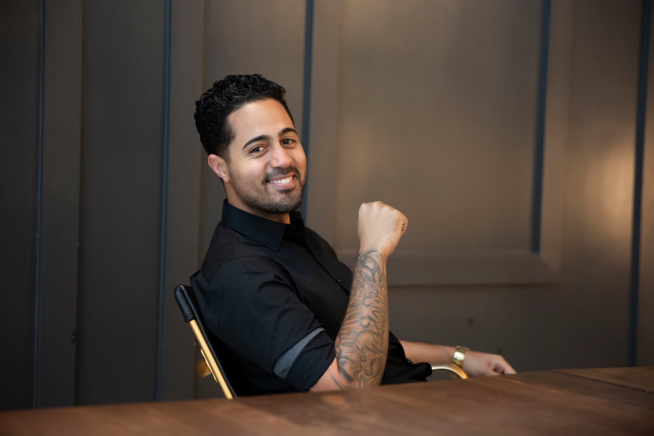 Smiling man in a black shirt seated at a wooden table, tattooed forearm raised.