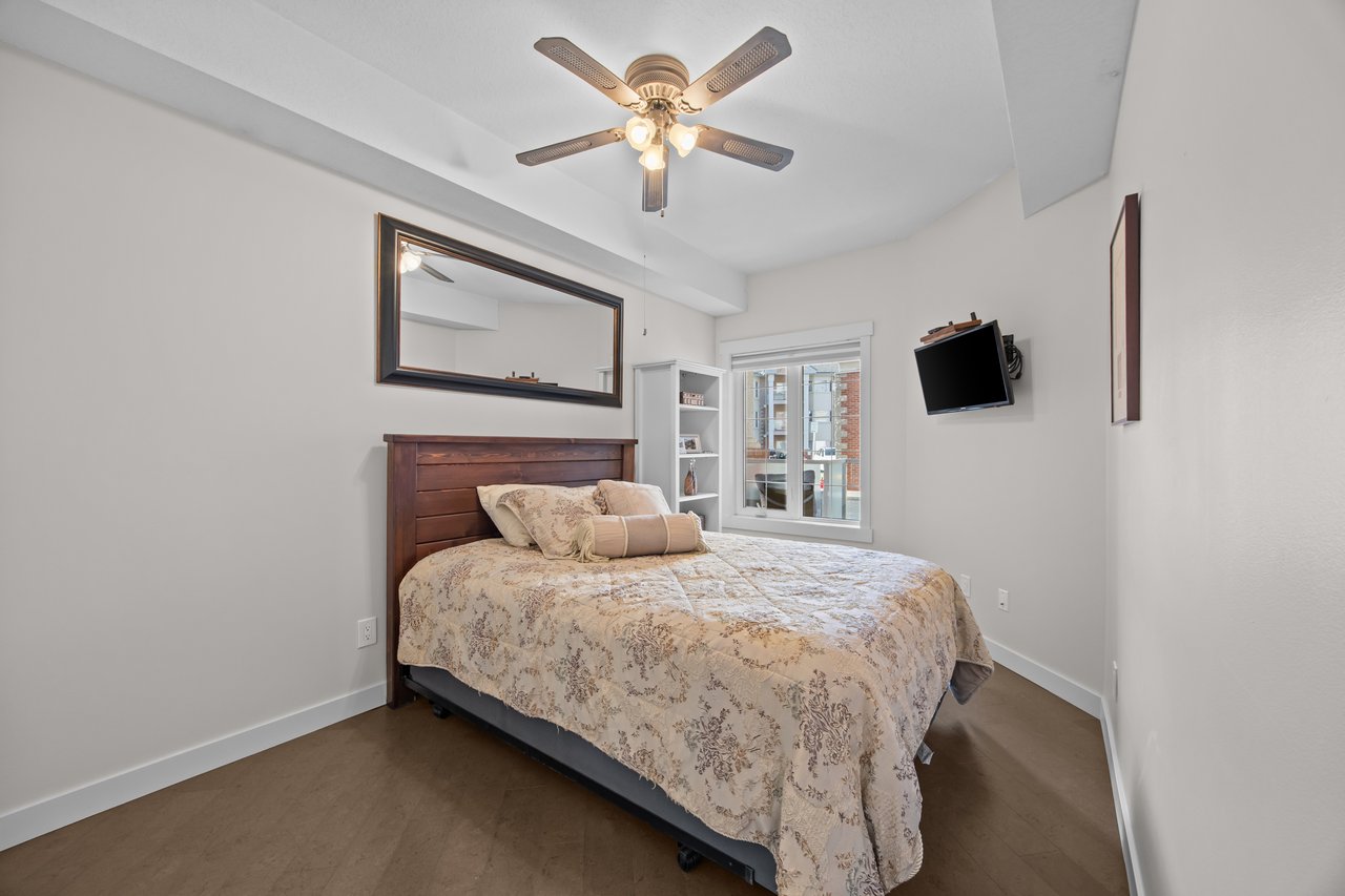 Furnished bedroom with a floral bedspread, ceiling fan, and window overlooking nearby buildings.