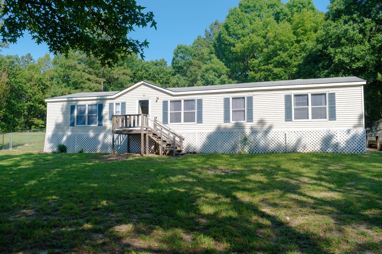 White manufactured home with blue shutters, a raised wooden porch, and lattice skirting on a tree-lined lawn.