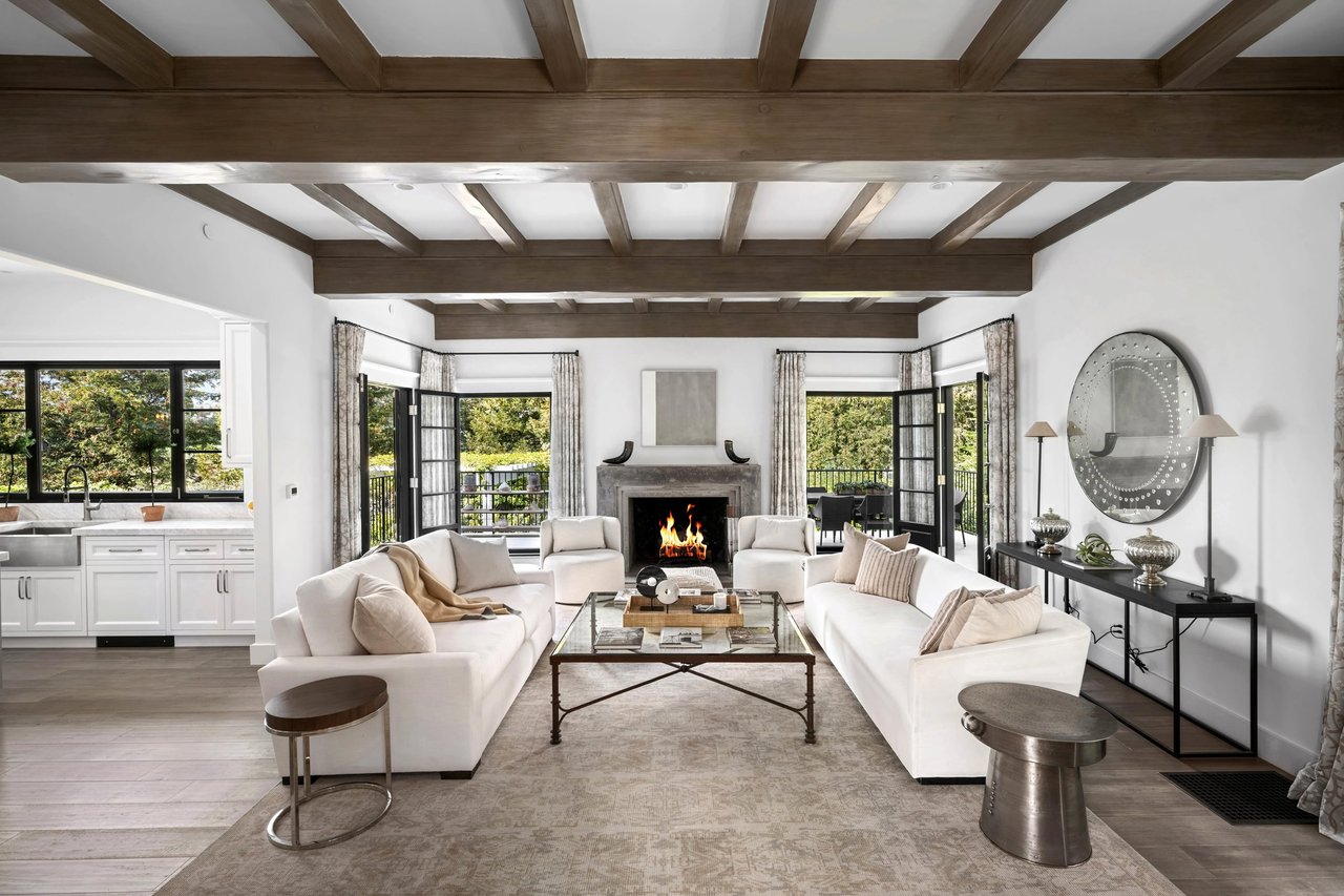Living room with exposed ceiling beams, a lit fireplace, white sofas, and doors opening to greenery.