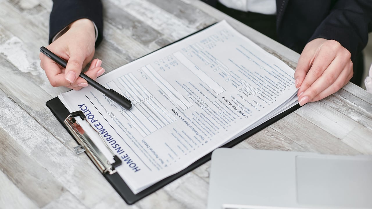 Hands completing a homeowners insurance policy form on a clipboard beside a laptop.