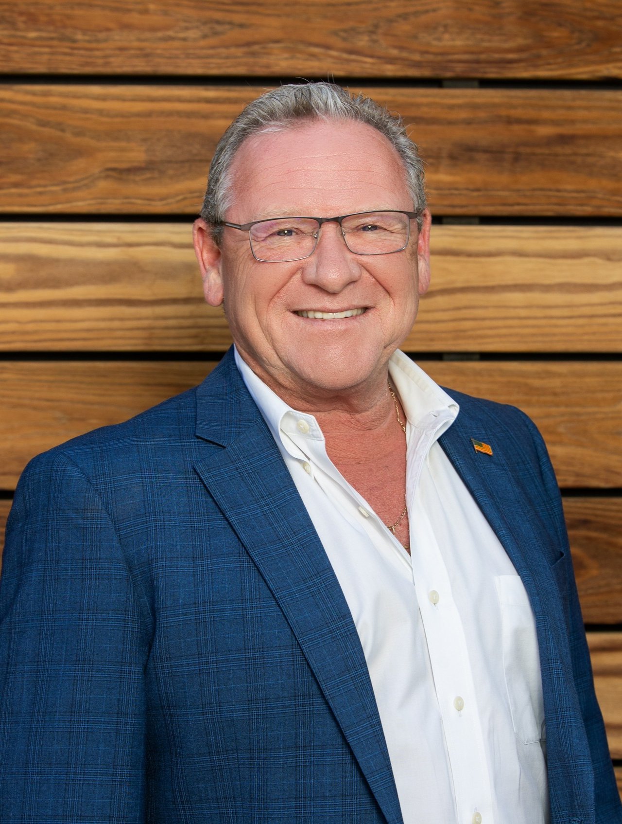 Johnny Walker smiling in a blue checked suit jacket and white shirt against a wood-plank wall.