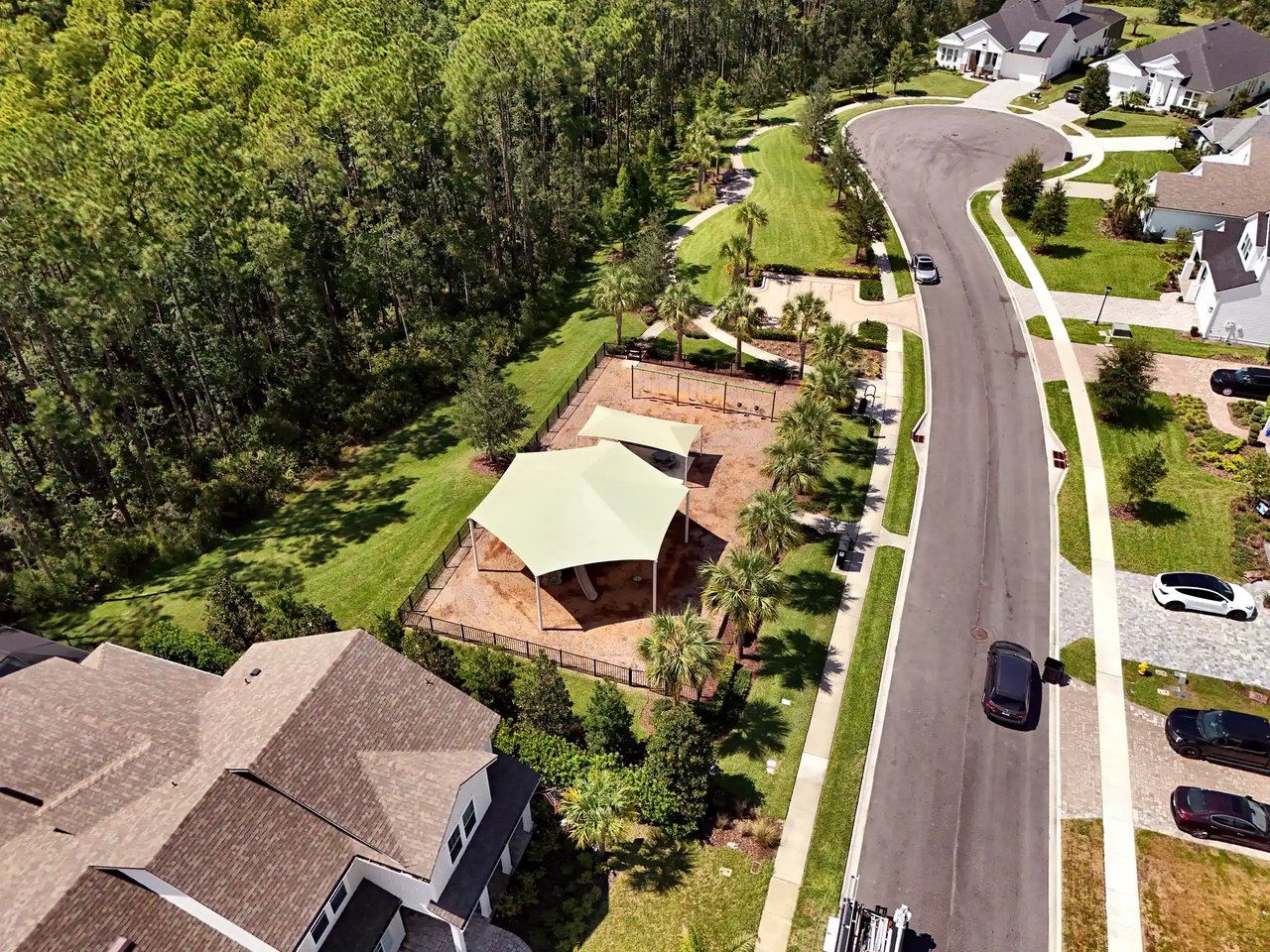 Aerial view of a fenced playground with shade canopies and palms beside a Palm Crest street, backed by woods.