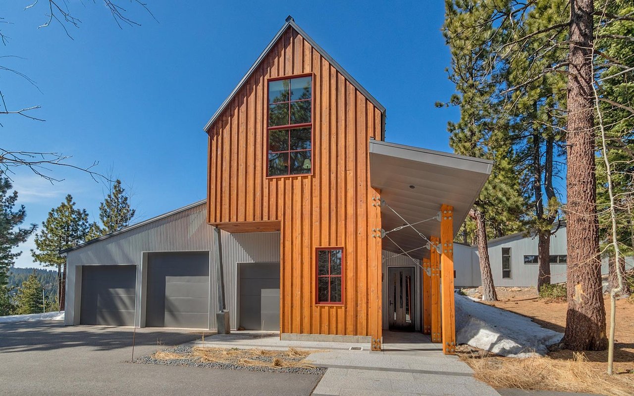 Modern home with a tall timber entry tower and three garage doors among pine trees and patches of snow.