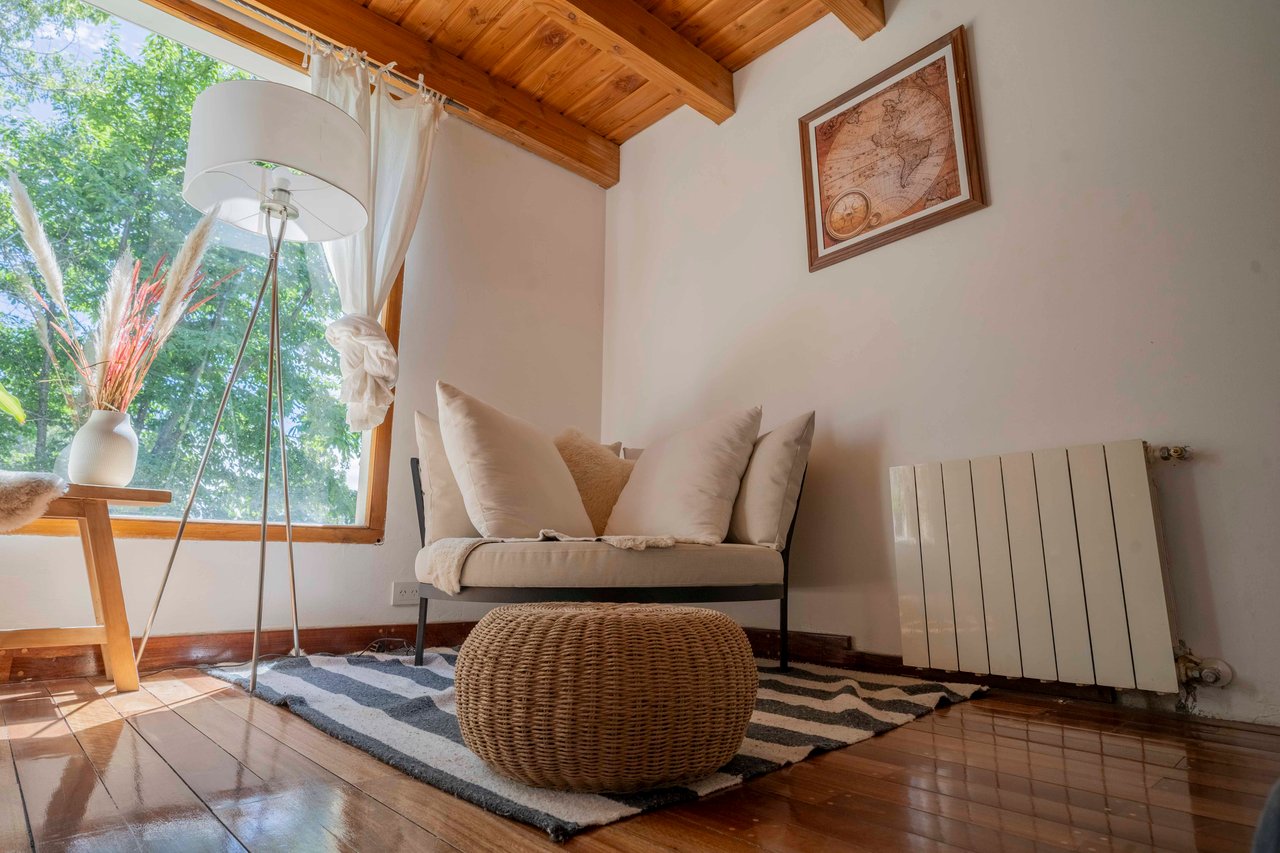Hotel sitting nook with a cushioned chair, woven ottoman, and large window beneath a wooden ceiling.