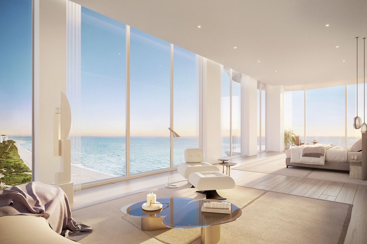 Oceanfront living room with floor-to-ceiling windows, pale furnishings, and sculptural lounge chairs.