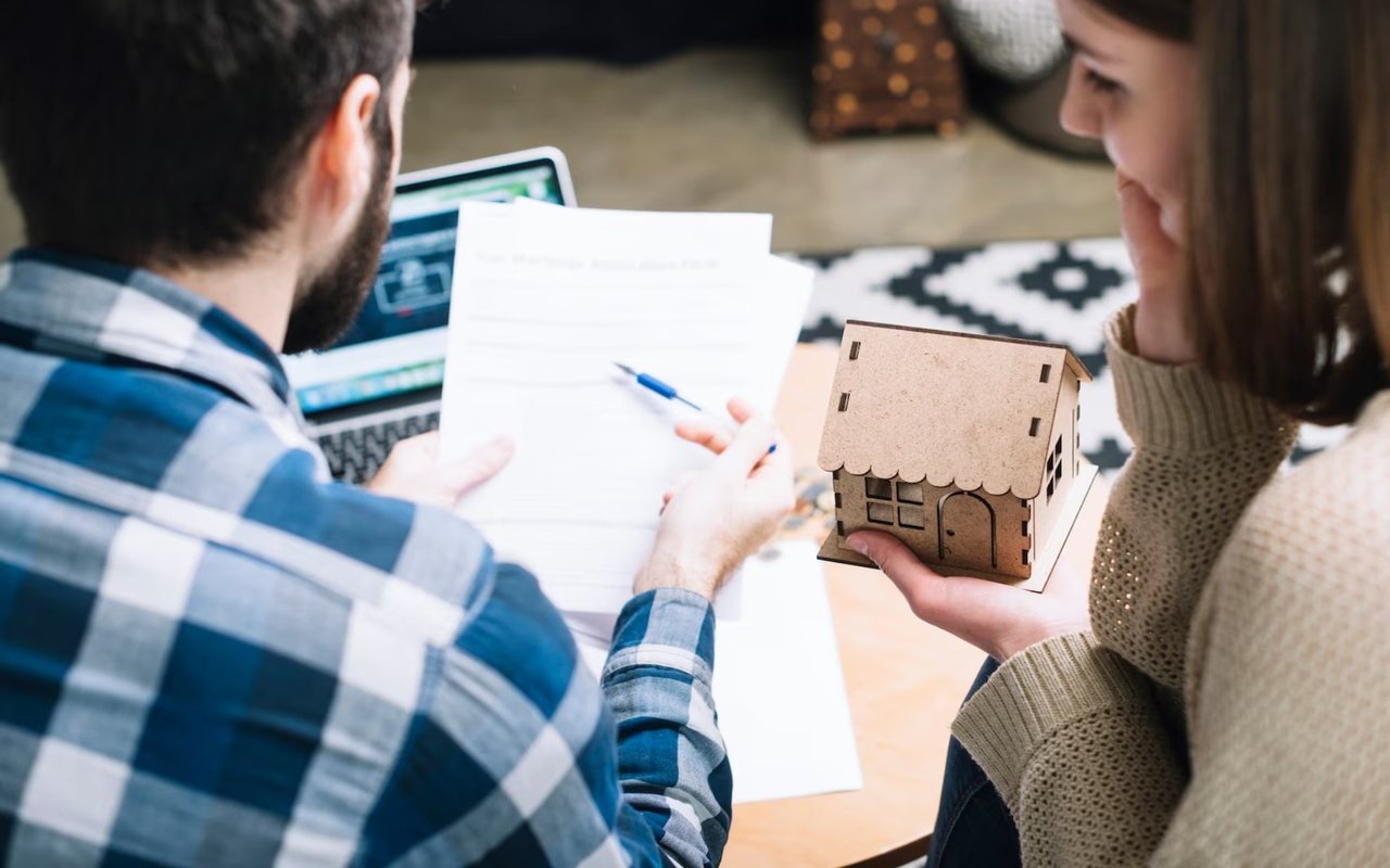 Two people review paperwork beside a laptop as one holds a small wooden model house.