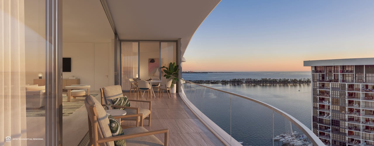 Furnished high-rise balcony with curved glass railing overlooking the water at sunset.