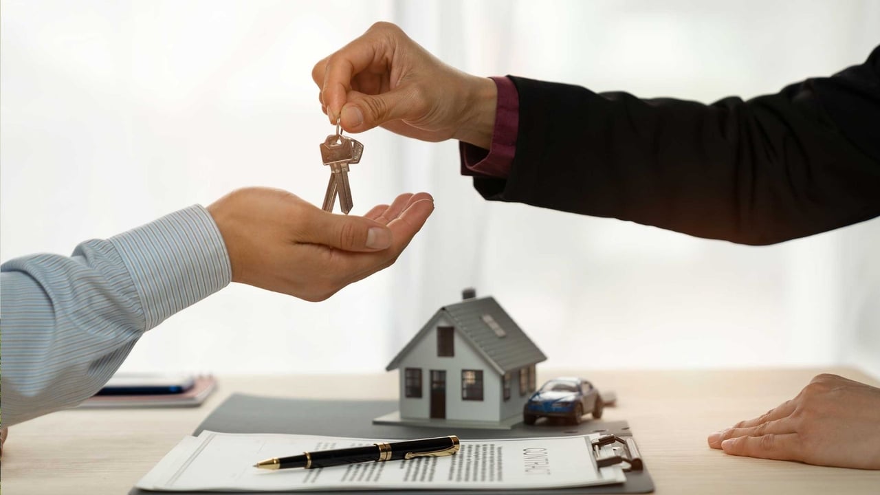 Hands exchange house keys above a contract, with a miniature home and car on the desk.