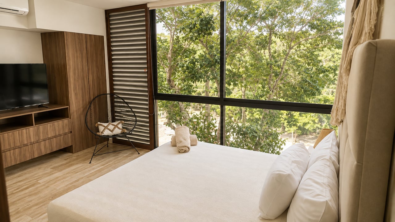 Bedroom with a large bed and floor-to-ceiling windows overlooking leafy trees.