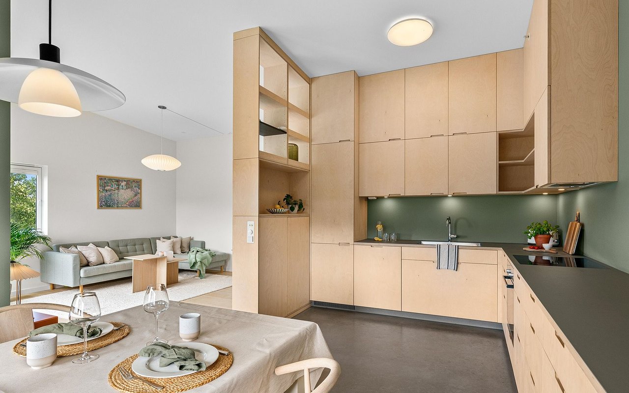 Open-plan kitchen with pale wood cabinets and green backsplash beside a furnished living area.