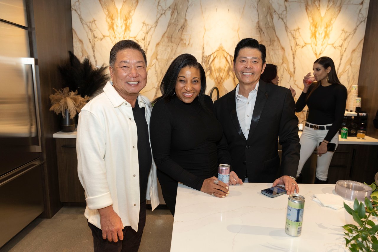 Three guests pose together at an open house beside a white counter and marble-patterned wall.