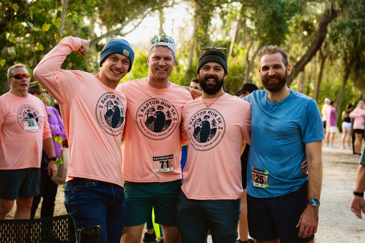 Four Raptor Run 5K participants pose on a wooded trail, three wearing pink event shirts.