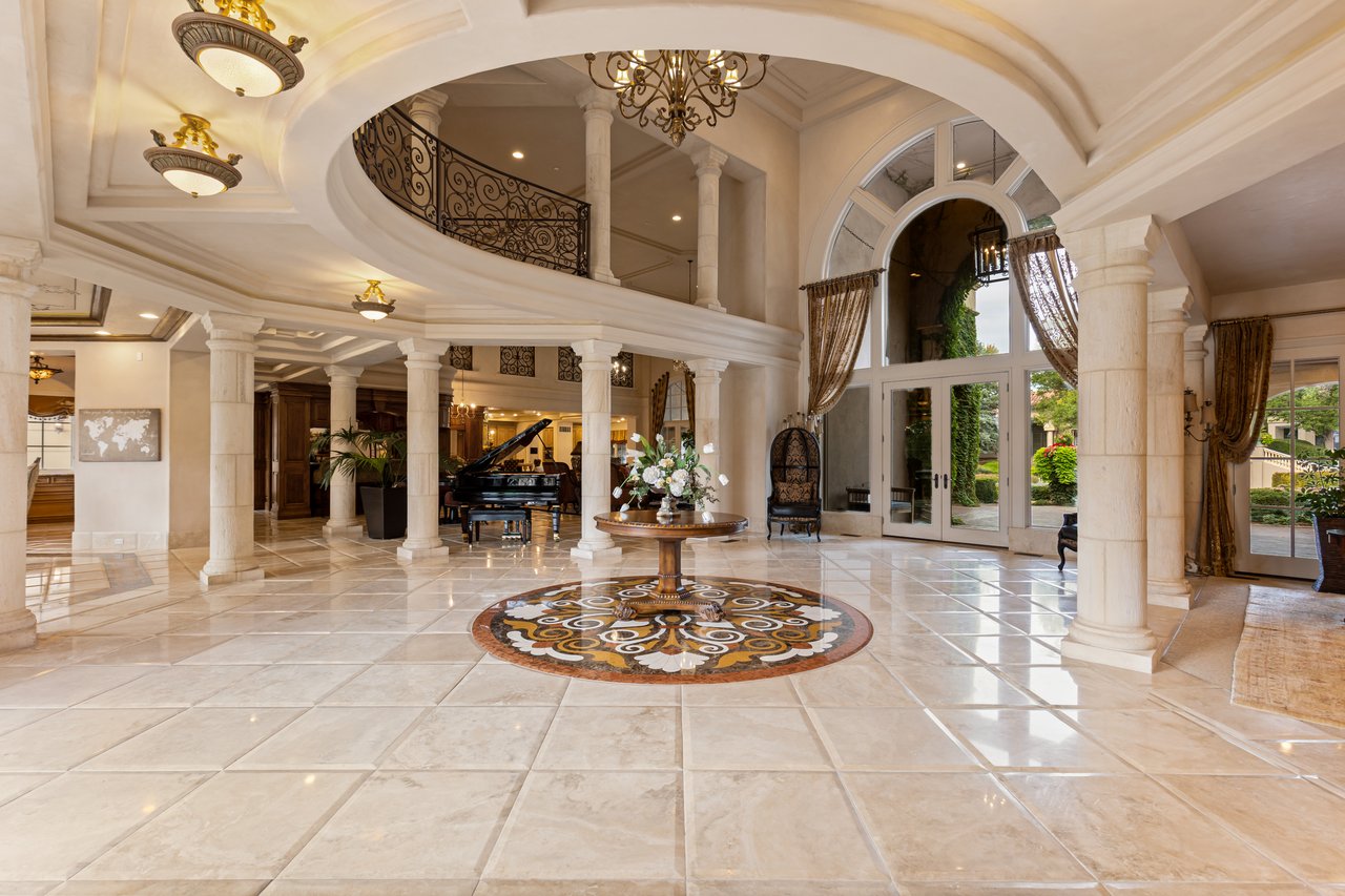 Grand foyer with marble floors, stone columns, arched glass doors, and a curved wrought-iron balcony.