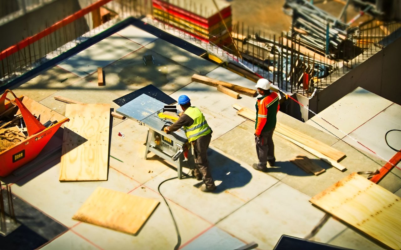 Construction workers cut plywood on a building site amid lumber and exposed rebar.