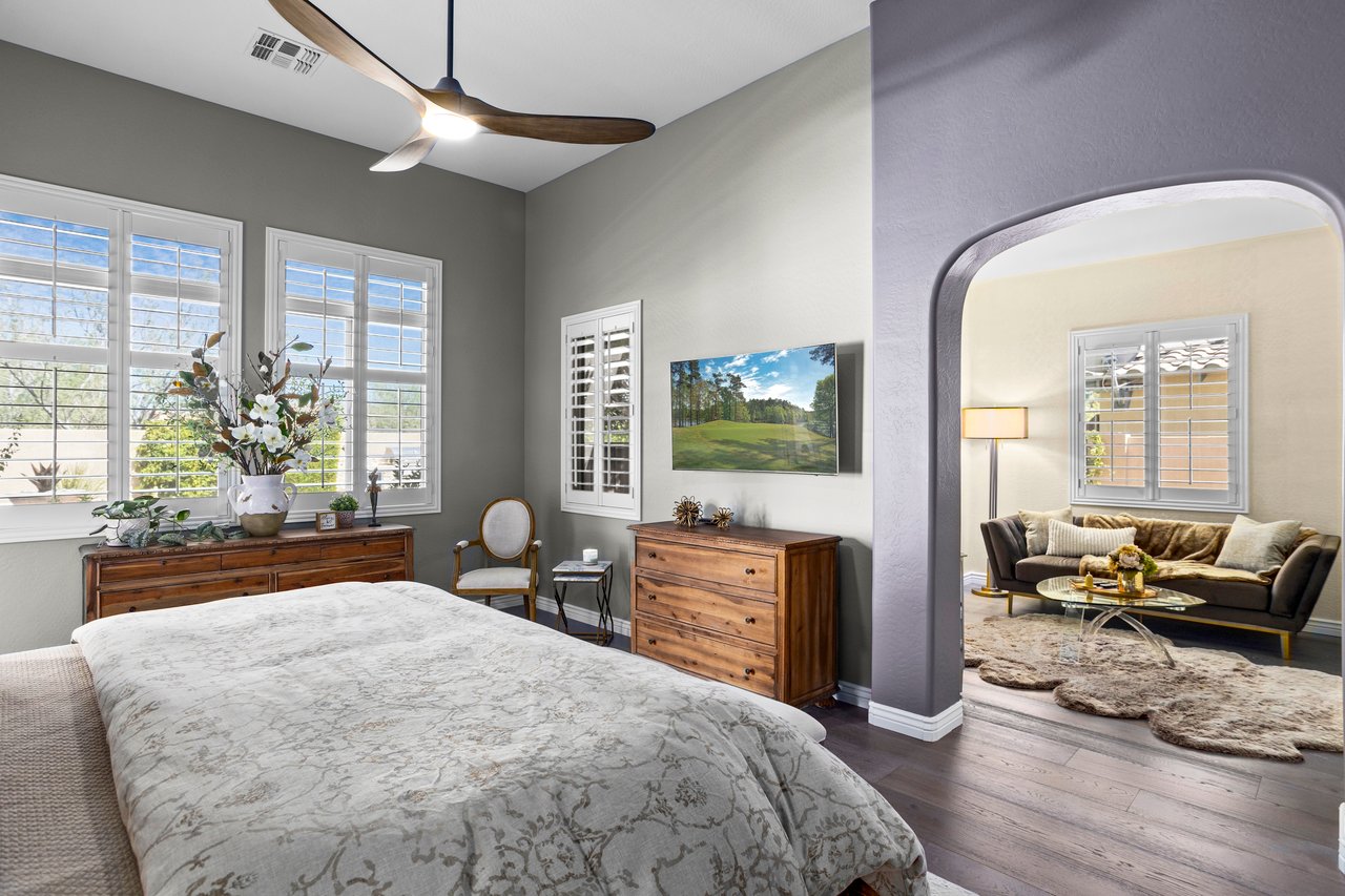 A staged bedroom with a bed in the foreground, windows with plantation shutters, wood dressers, and a sitting area visible through an arched opening.