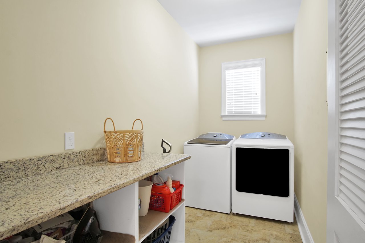 Laundry room with beige walls featuring a granite countertop with a wicker basket, white washer and dryer, under-shelf storage, and a window. Clean and organized.