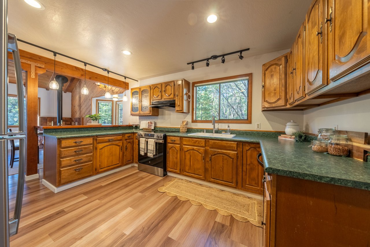 Kitchen with warm wood cabinetry, green countertops, and windows overlooking trees.