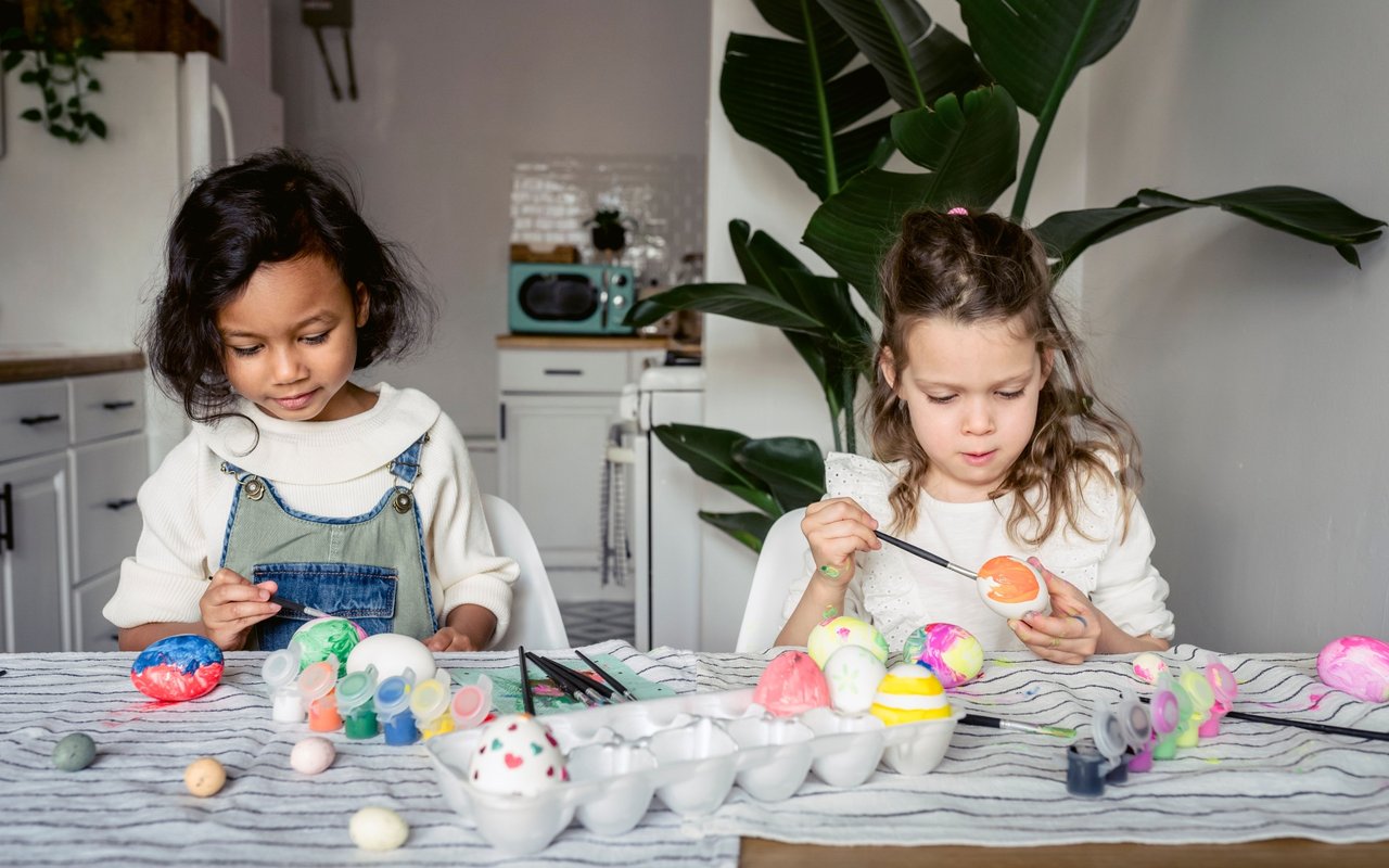 Two children decorate Easter eggs with paint and brushes at a kitchen table.