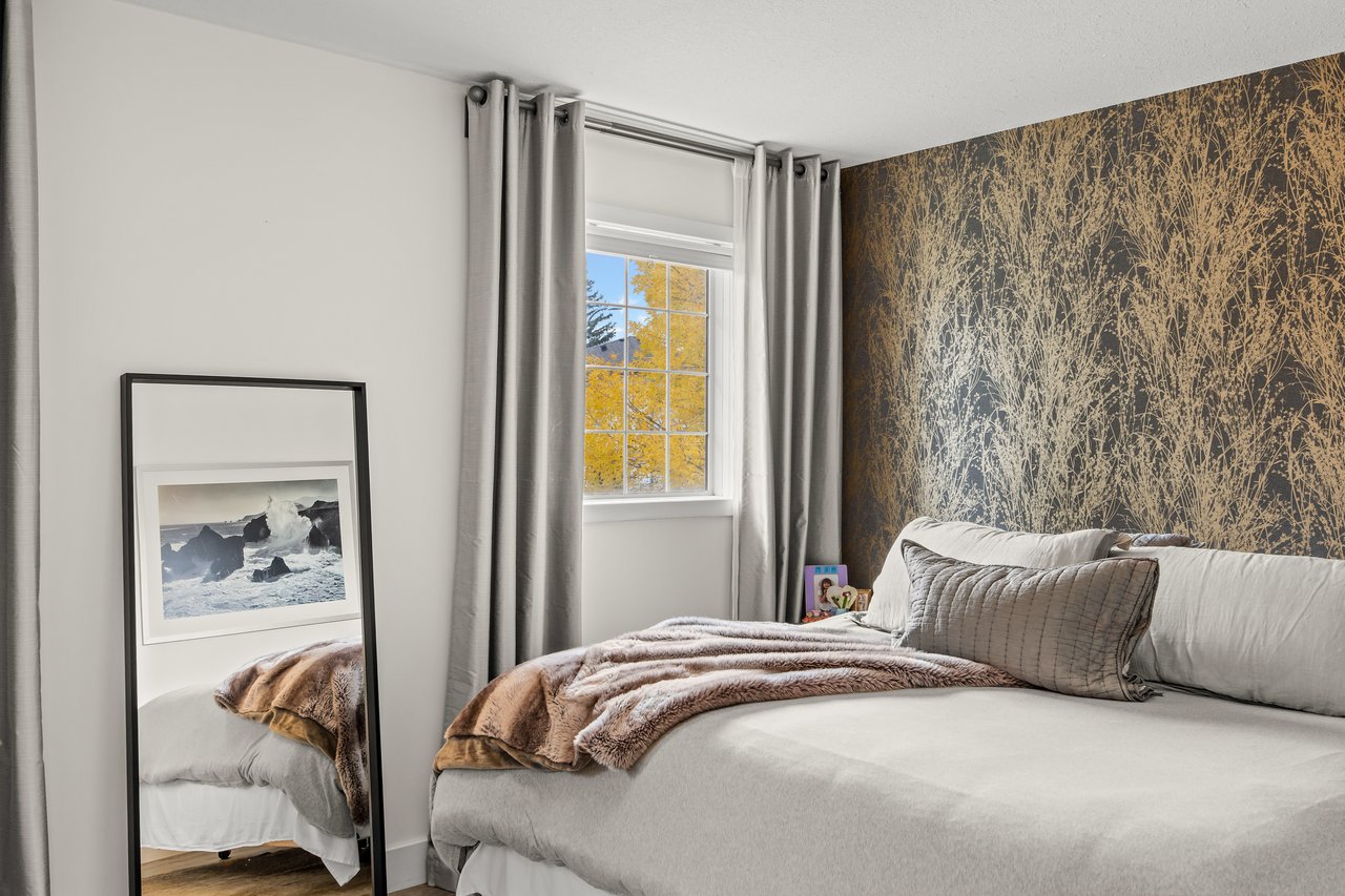 Furnished bedroom with a botanical accent wall, layered bedding, and a window overlooking autumn foliage.