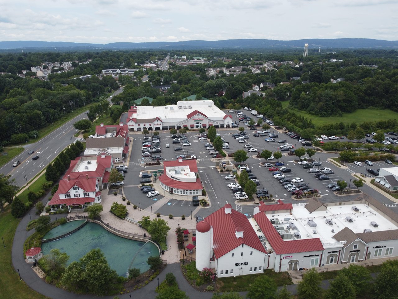 High-altitude aerial drone view highlighting the red-roof commercial buildings, silo landmark, and surrounding landscape at Purcellville Gateway in Purcellville, VA, captured by local real estate expert Diane Geremia.