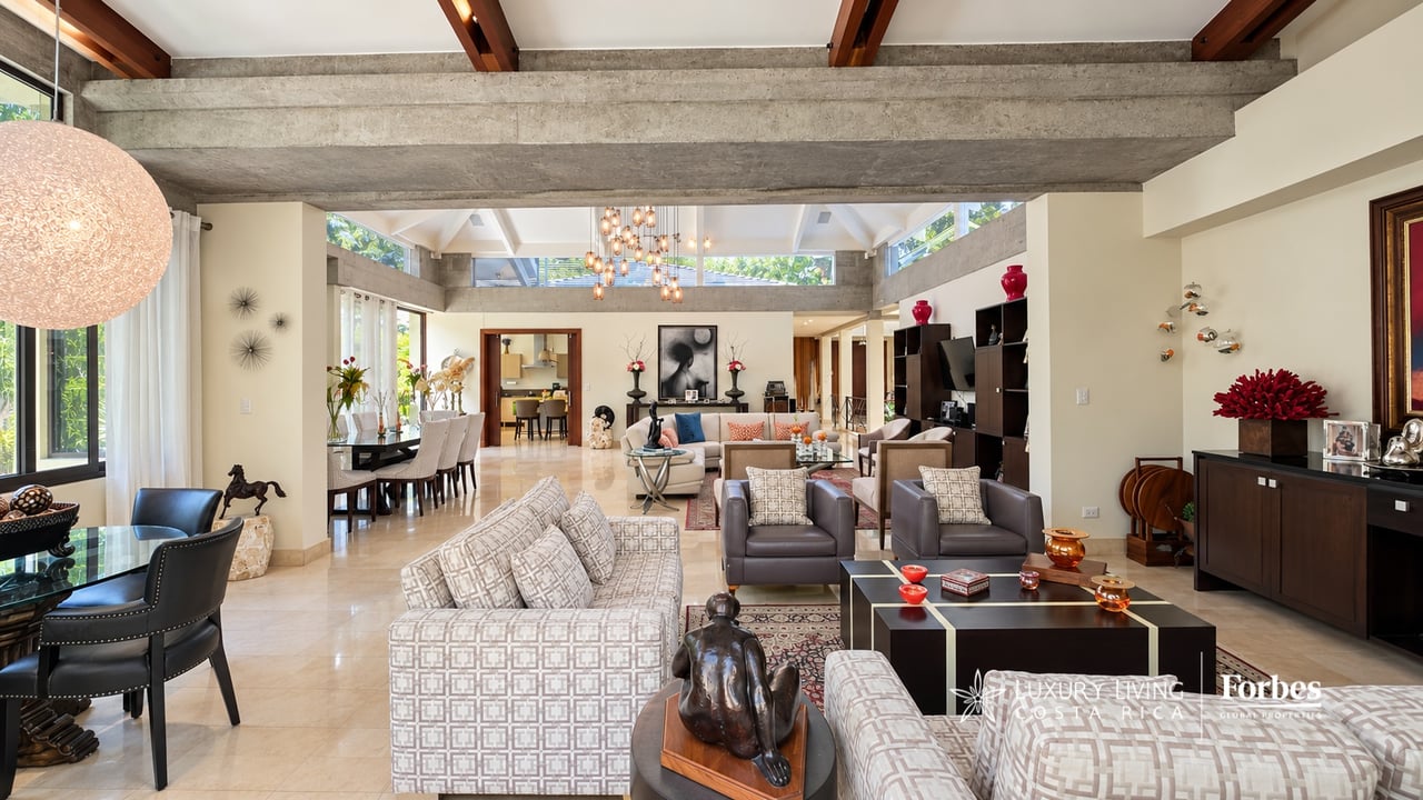 Living room with exposed concrete beams, patterned sofas, and large windows overlooking greenery.