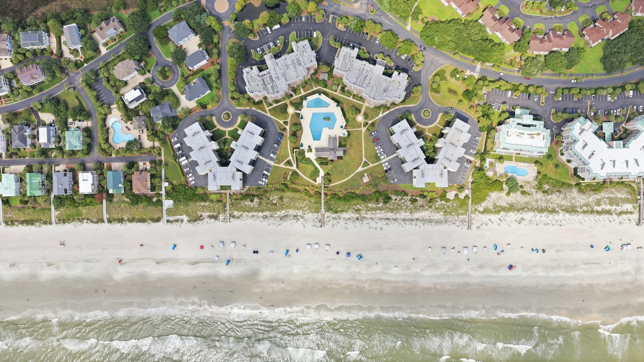 Beachfront villas and swimming pools beside a wide sandy beach and ocean, seen from above.