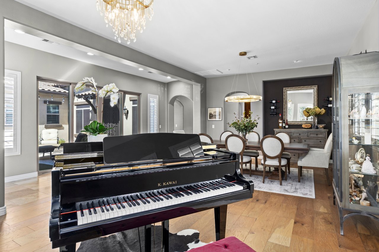 A furnished dining room with a grand piano, chandelier, wood floors, and a dark accent wall with a sideboard and mirror.