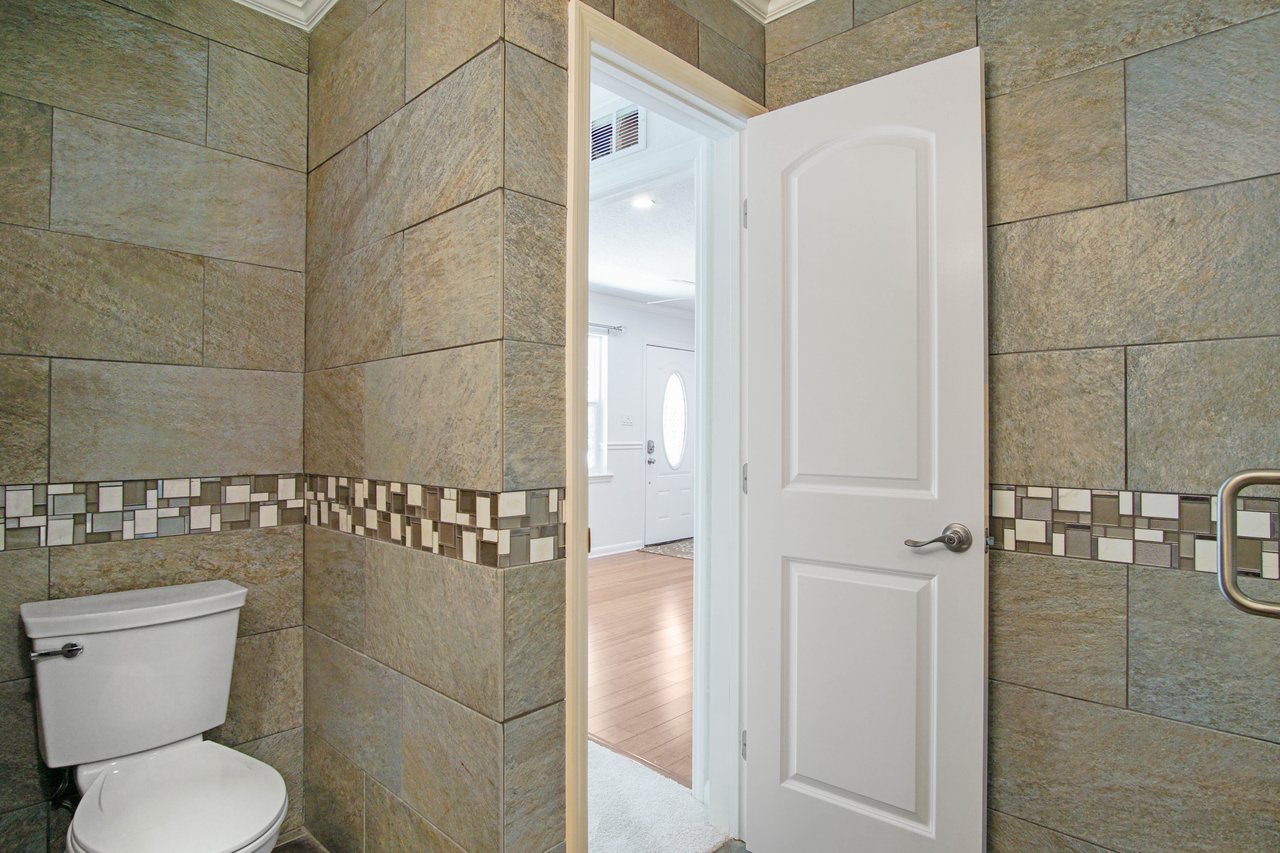 Bathroom with dark tiled walls and white door slightly open, revealing a hallway with hardwood floors and an oval glass door. Calm, tidy atmosphere.
