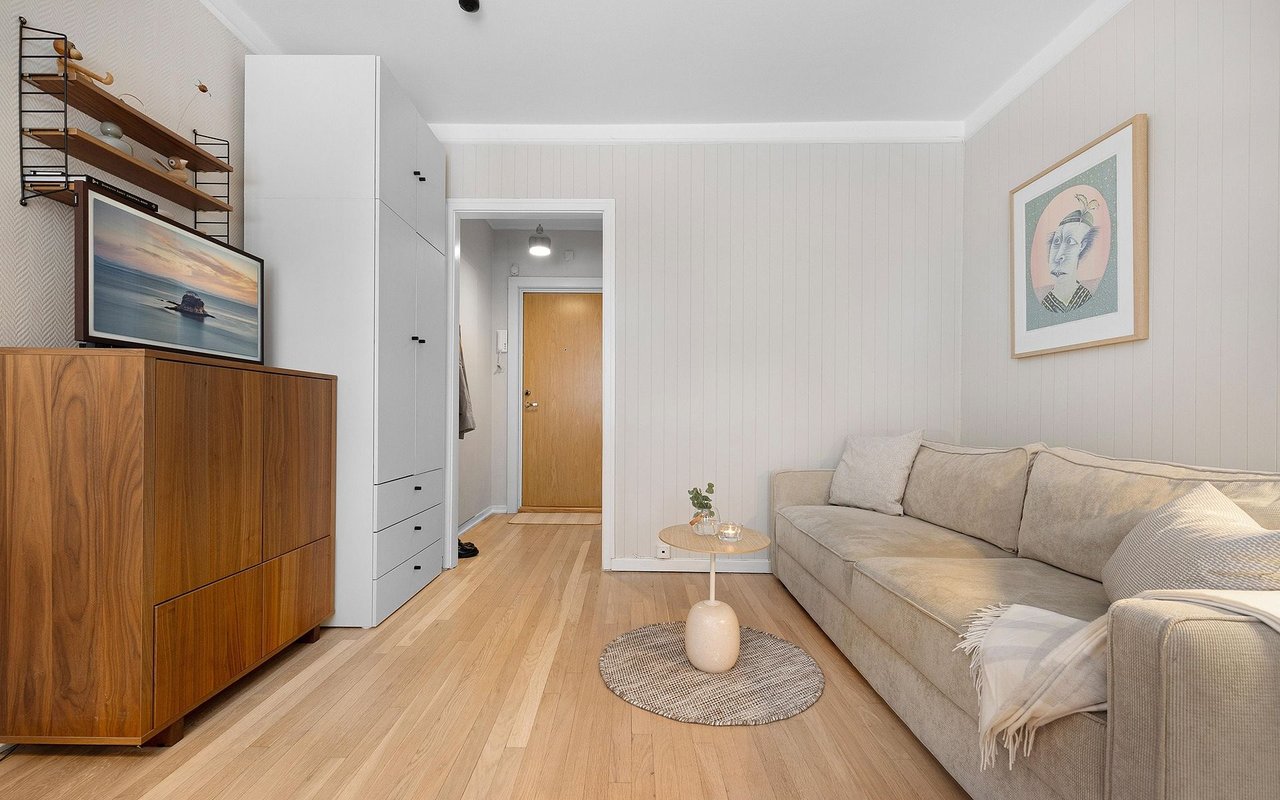 Living room with a beige sectional, pale wood floors, and a walnut cabinet beneath wall shelving.