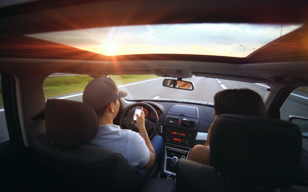 Two people drive along a highway at sunset, viewed from the back seat through a sunroof.