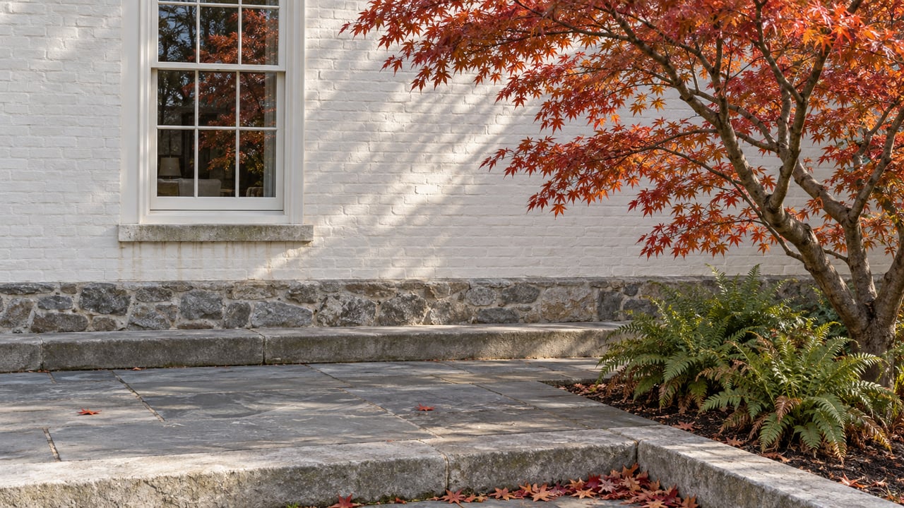 Autumn maple beside a white brick home's garden terrace, with a tall sash window and weathered blue-gray stone paving.