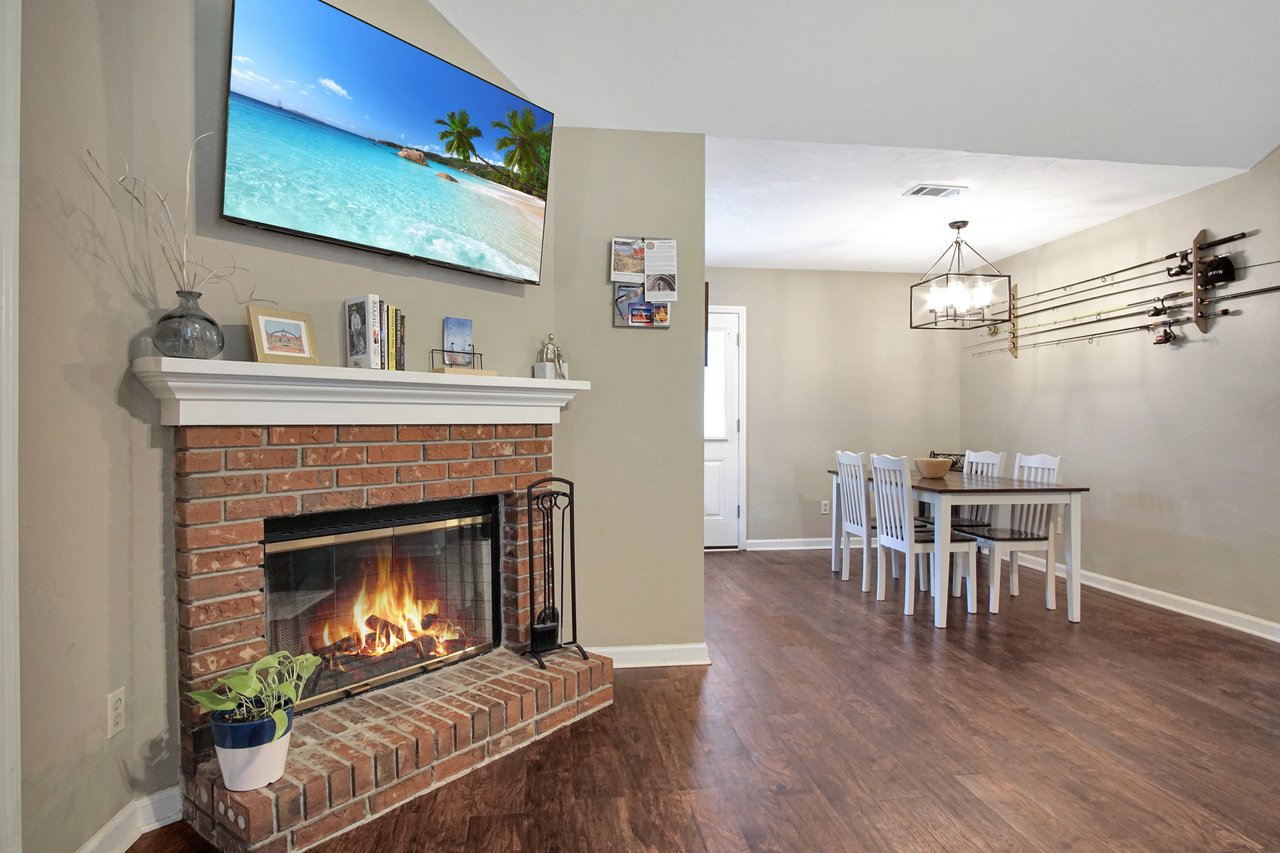 A cozy room with a lit brick fireplace, a tropical beach image on the TV above, a white mantle with books and decor, and a dining table with white chairs. Fishing rods adorn the opposite wall, and the wood floor adds warmth to the inviting atmosphere.