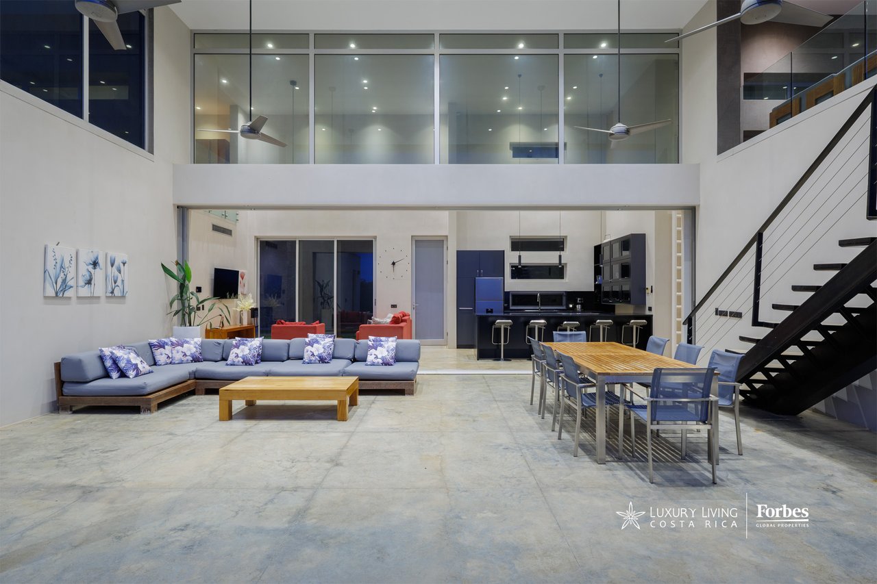Double-height living and dining area with a gray sectional, dark kitchen, and open staircase.