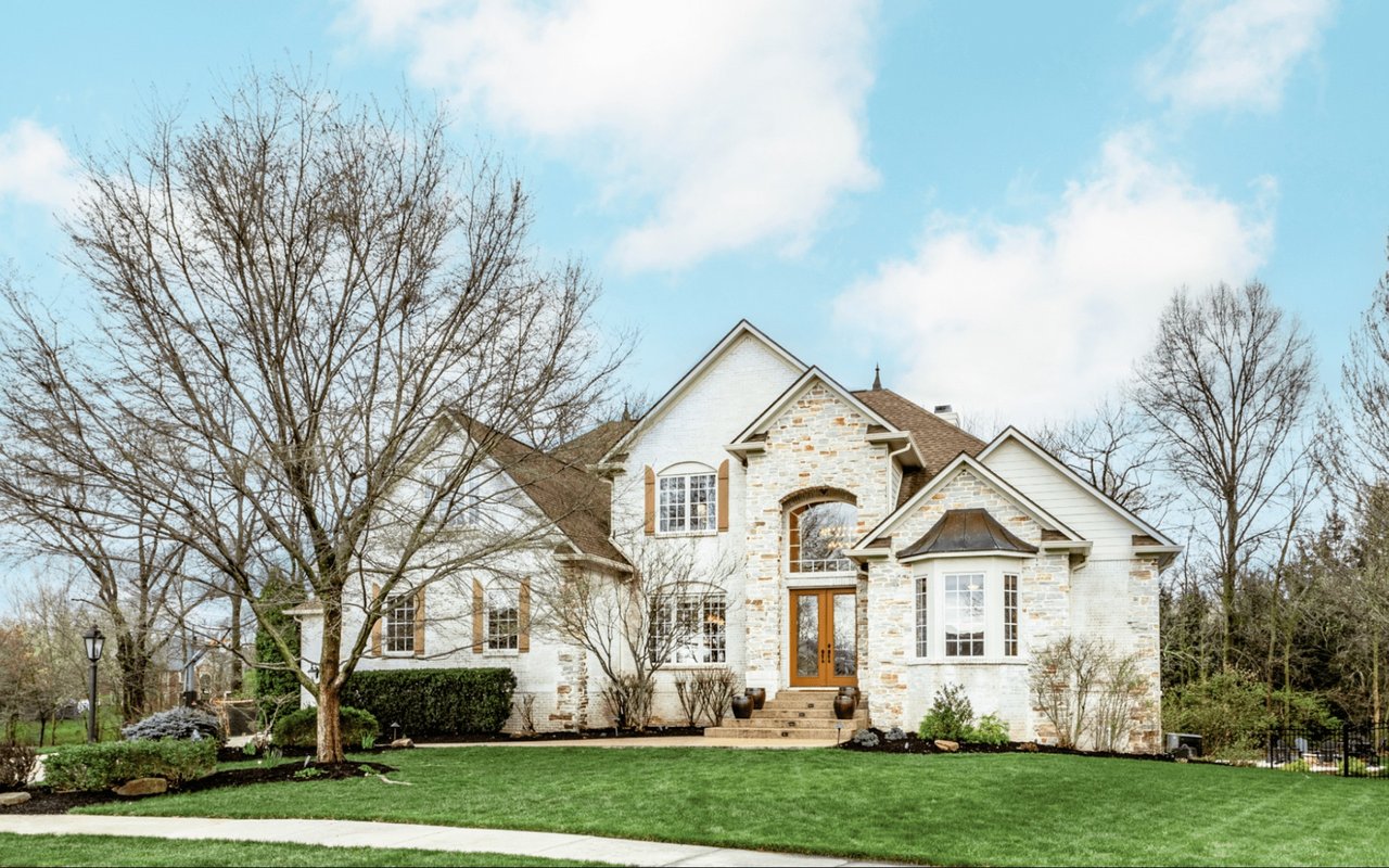An elegant, traditional-style luxury home displaying high curb appeal, a stone-accented facade, custom arched entry, multi-pane windows, and manicured landscaping with a paved walkway.