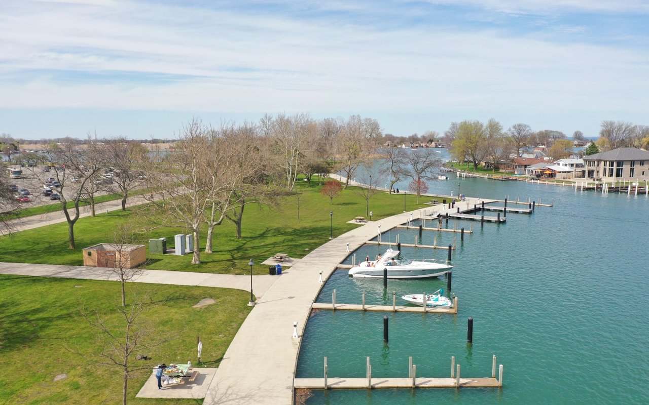 Waterfront marina with boats at docks beside a curving park promenade and green lawn.
