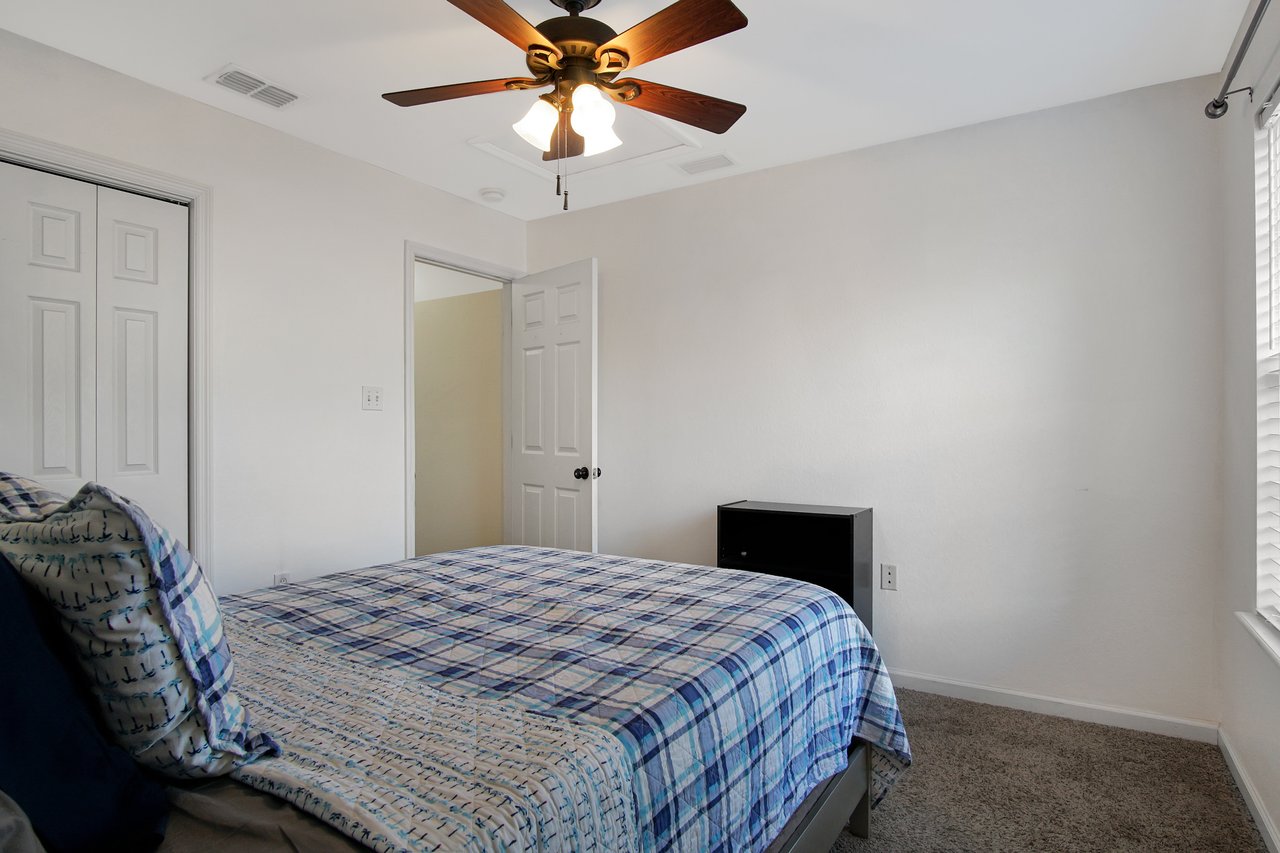 Bedroom with a plaid blue bedspread, pillows, a ceiling fan, and closed white closet doors. A small black nightstand is beside the bed. Natural light from a window illuminates the space.