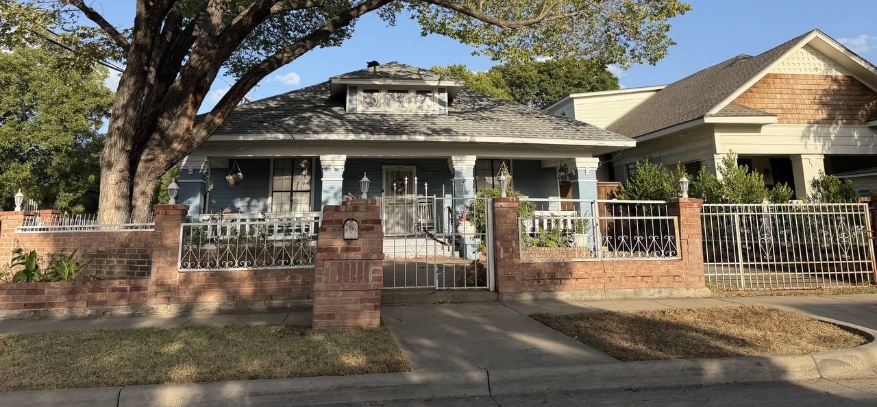 Blue-gray house behind a brick wall and decorative metal fence beneath a large shade tree.