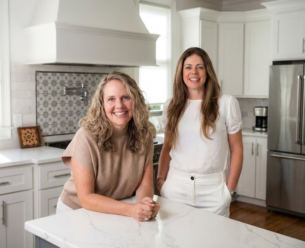 Two women pose in a bright kitchen, leaning against a marble island.