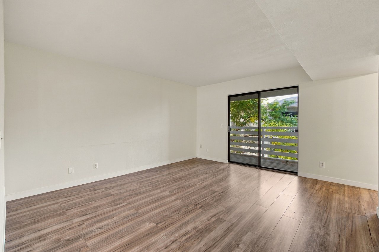 Empty living room with wood-look flooring and a sliding glass door to a tree-lined balcony.