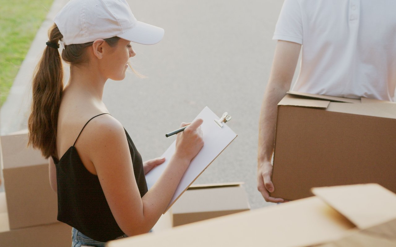 A woman checks a clipboard beside moving boxes as another person carries a box.