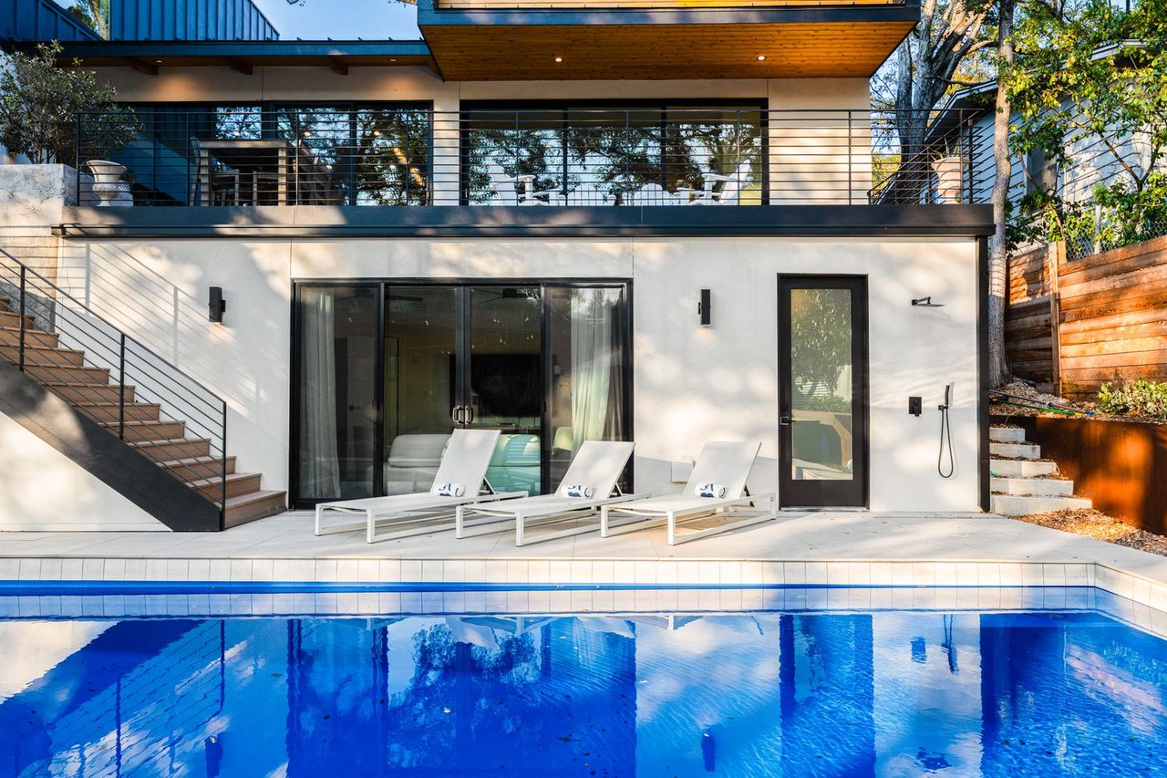 Modern two-story home with a blue pool, three patio loungers, and exterior stairs.